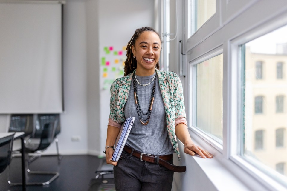 Woman wearing casuals holding laptop standing in office. Portrait a confident woman standing by the window in office and looking at camera.