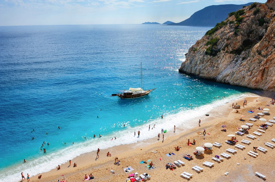 Aerial view of a Mediterranean beach in Fethiye, Turkey, with people swimming and sunbathing, and a sailboat in the bay.