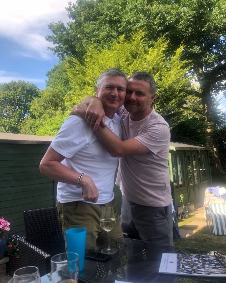 two men are posing for a picture in front of a green shed