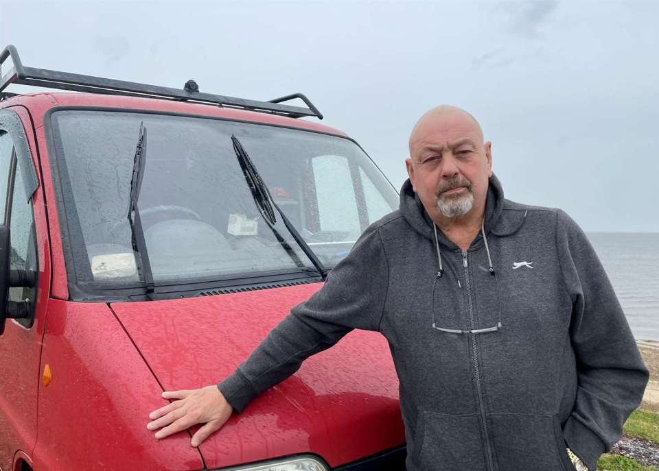 a man wearing a grey sweatshirt with a slazenger logo on it stands in front of a red van