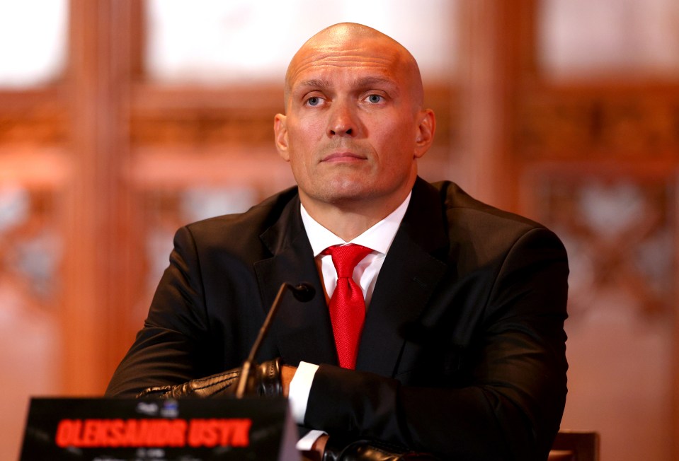 a bald man in a suit and tie sits in front of a sign that says aleksandr usyk