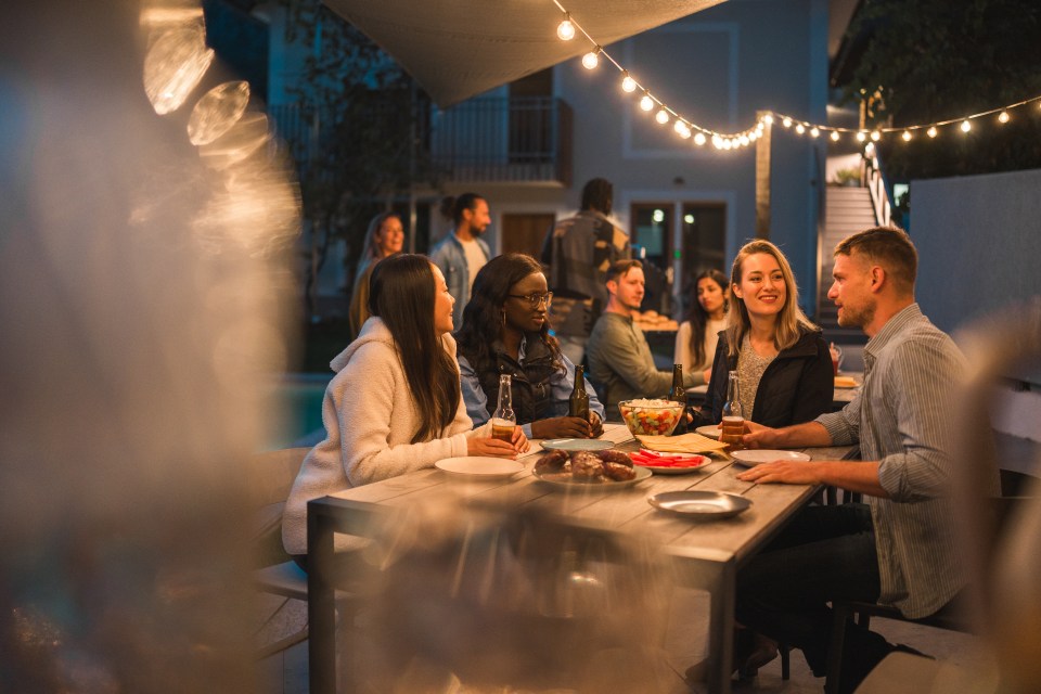 A mixed group of men and women engage in conversation over a meal at an evening outdoor party, with string lights adding ambiance to the diverse gathering.