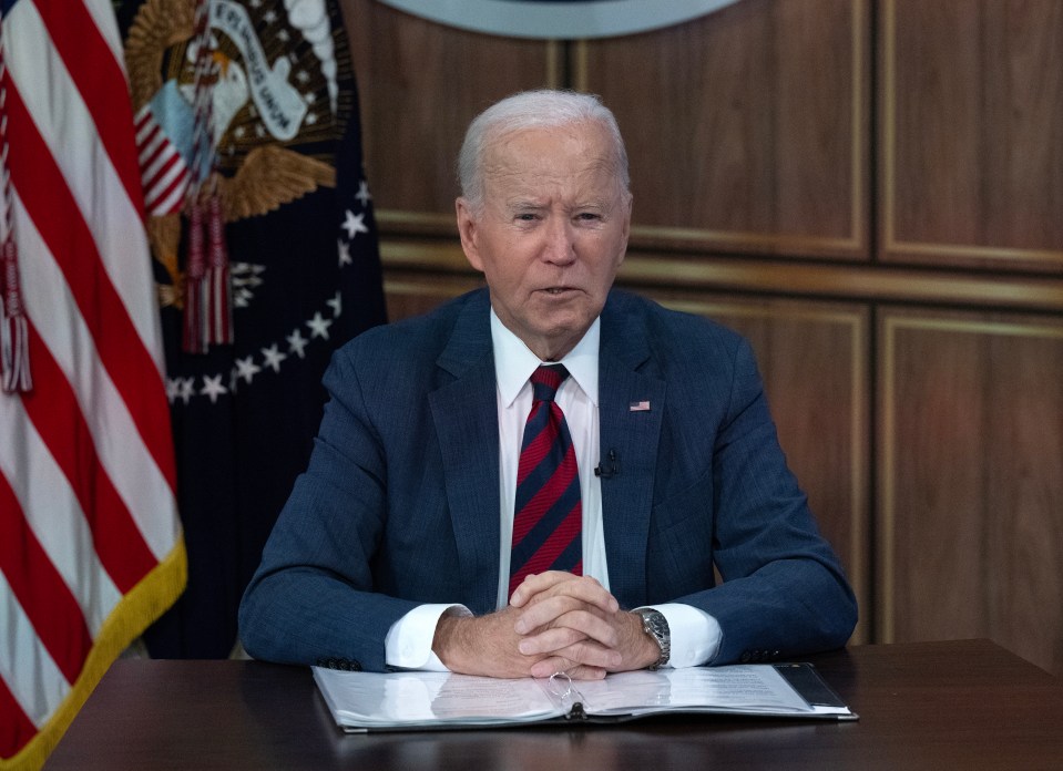 a man in a suit and tie sits at a desk in front of an american flag