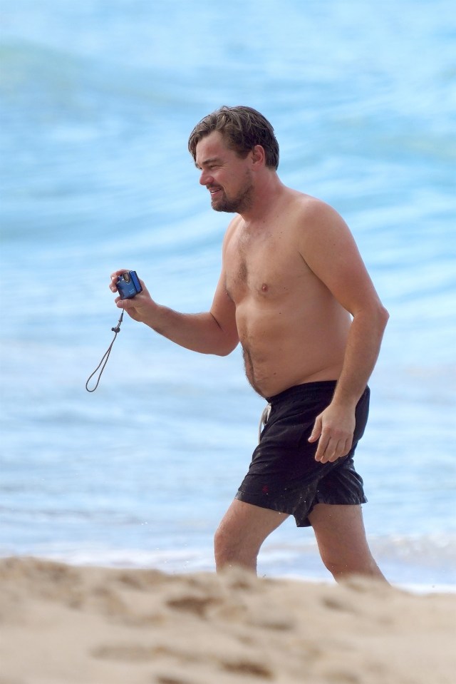 a shirtless man walking on the beach holding a camera
