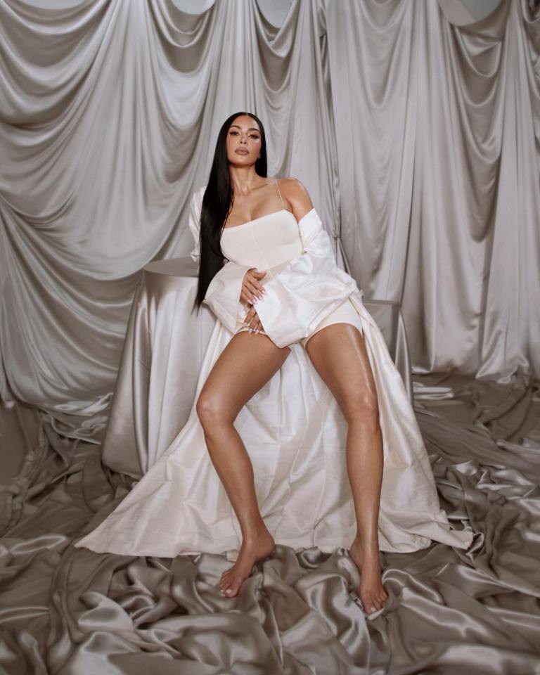 Kim Kardashian in a white bodysuit and robe, barefoot on silk fabric.
