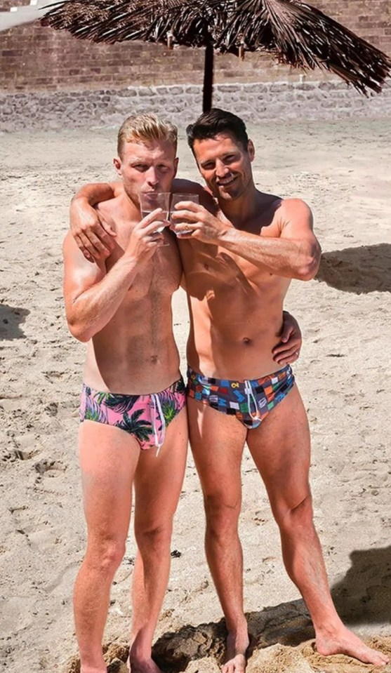Two men in swimsuits on a beach under a parasol, toasting drinks.