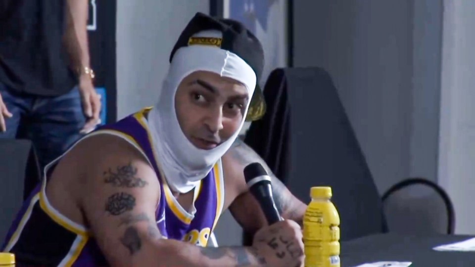 A man wearing a balaclava and Lakers jersey sits at a table holding a microphone.