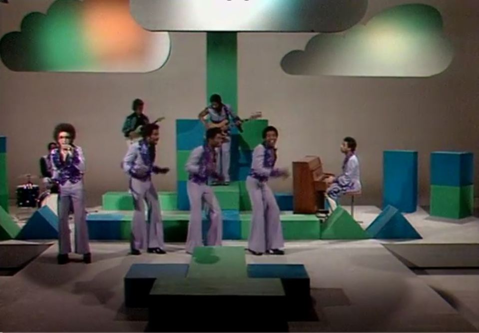 A band performs on a stage with colorful geometric blocks and cloud-shaped decorations.