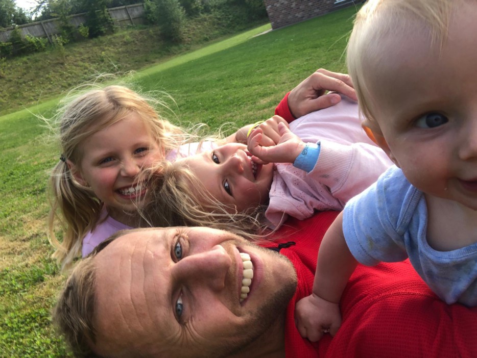 A man lying on the grass with three young children on top of him.
