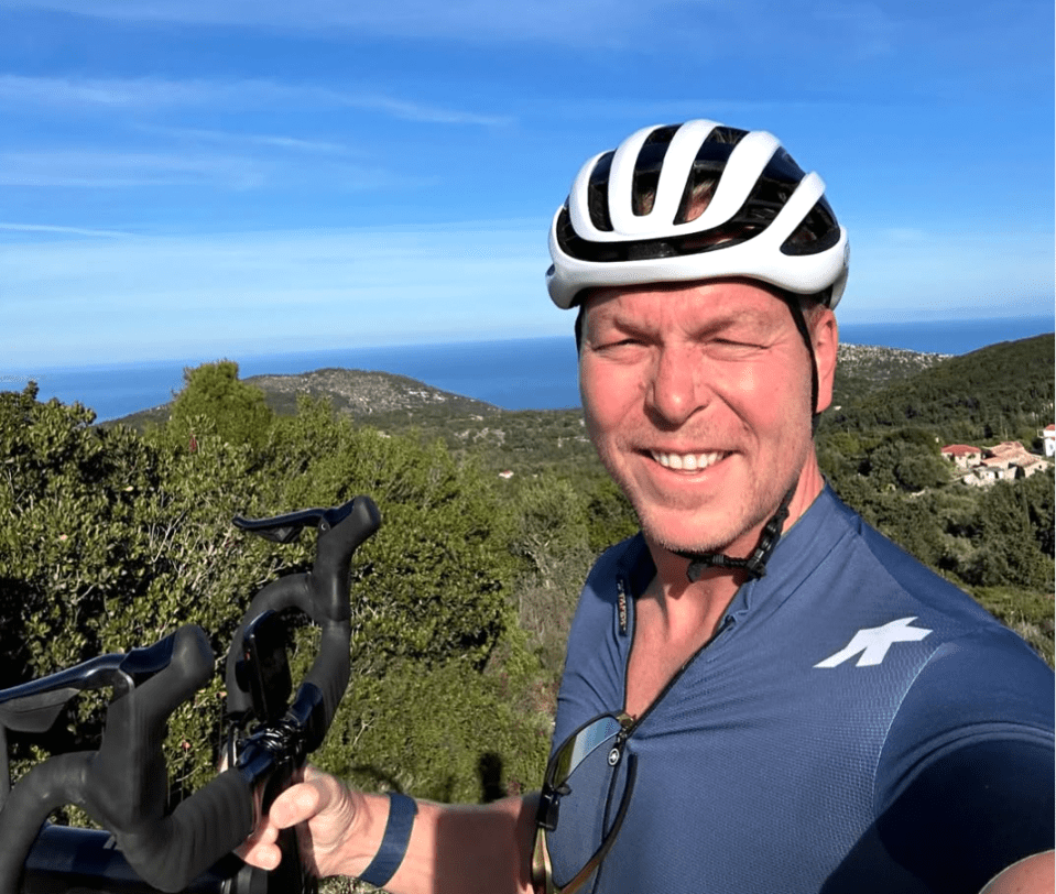 Man in cycling gear and helmet smiling at camera with bike handlebars in hand, overlooking ocean.