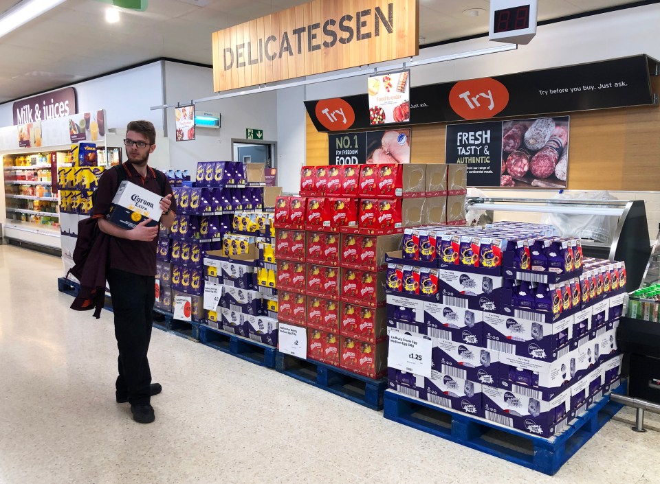 A man in a supermarket holds a box of Corona beer while Easter eggs are displayed on pallets nearby.