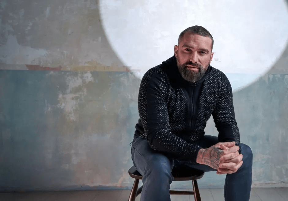 Portrait of a man with tattoos sitting on a stool.