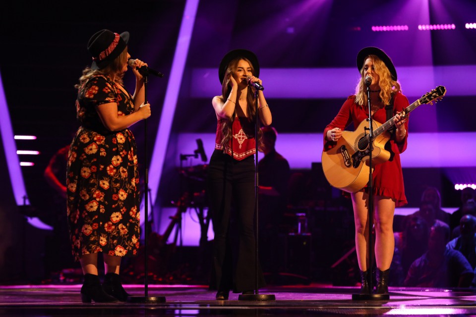 Three women singing on stage, one playing an acoustic guitar.