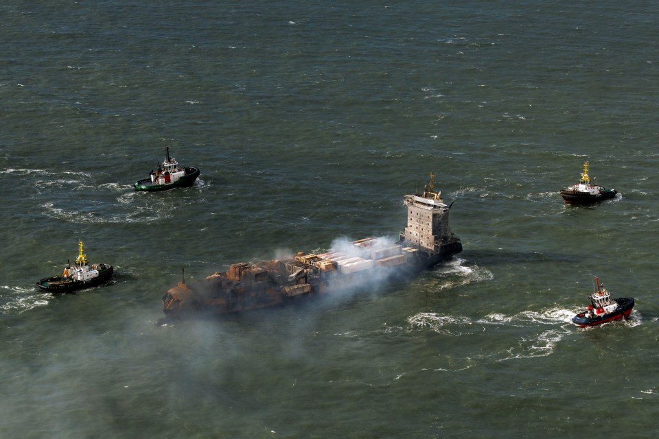 Aerial view of a cargo ship on fire in the sea, surrounded by tugboats.