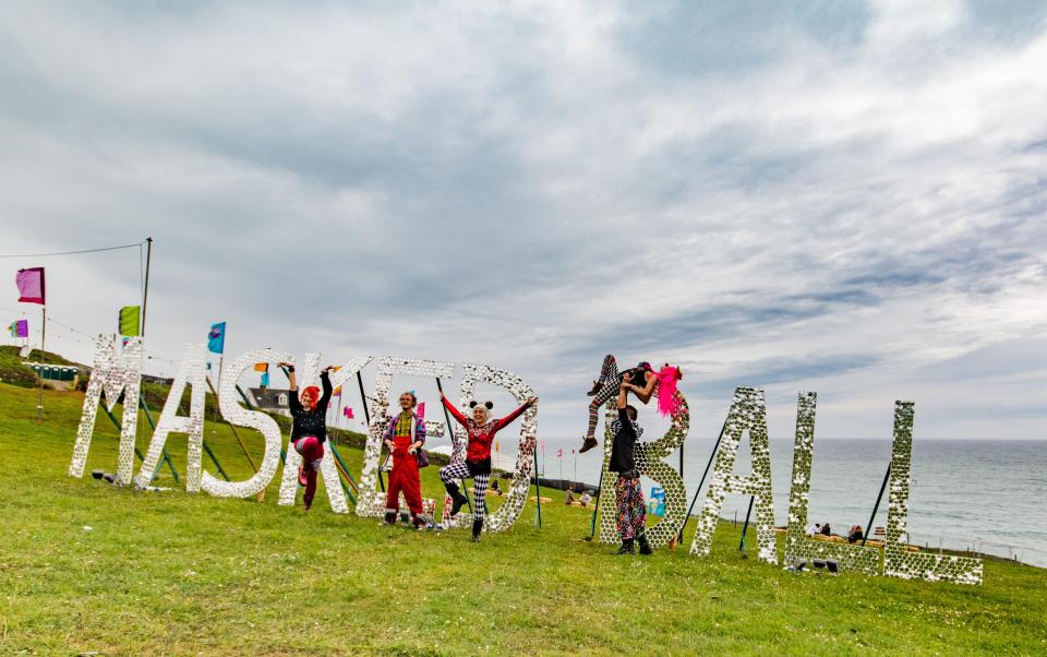 People posing with a large, mirrored "MASKERBALL" sign at a festival by the ocean.