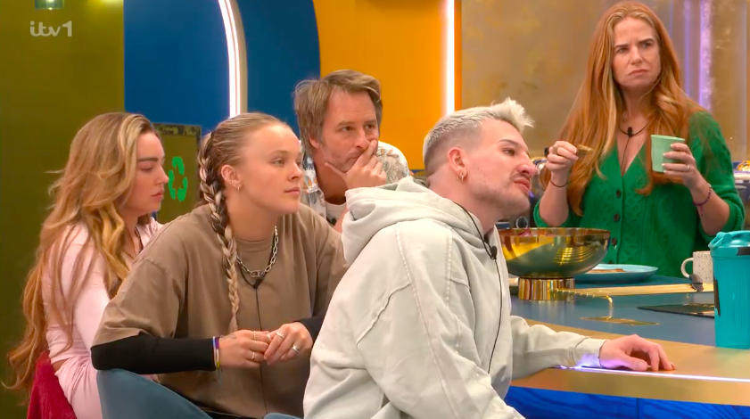 Screenshot of five people sitting at a table, one woman standing behind them.