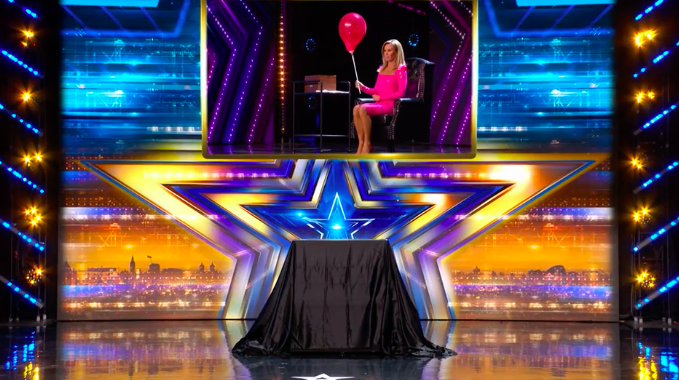 Screenshot of a woman in a pink dress holding a balloon on a stage with a covered box in the foreground.