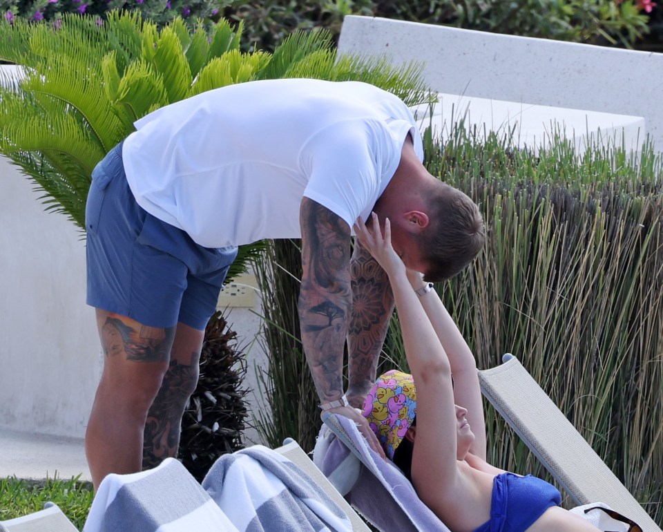 A man leans down to kiss a woman relaxing on a lounge chair by a pool.