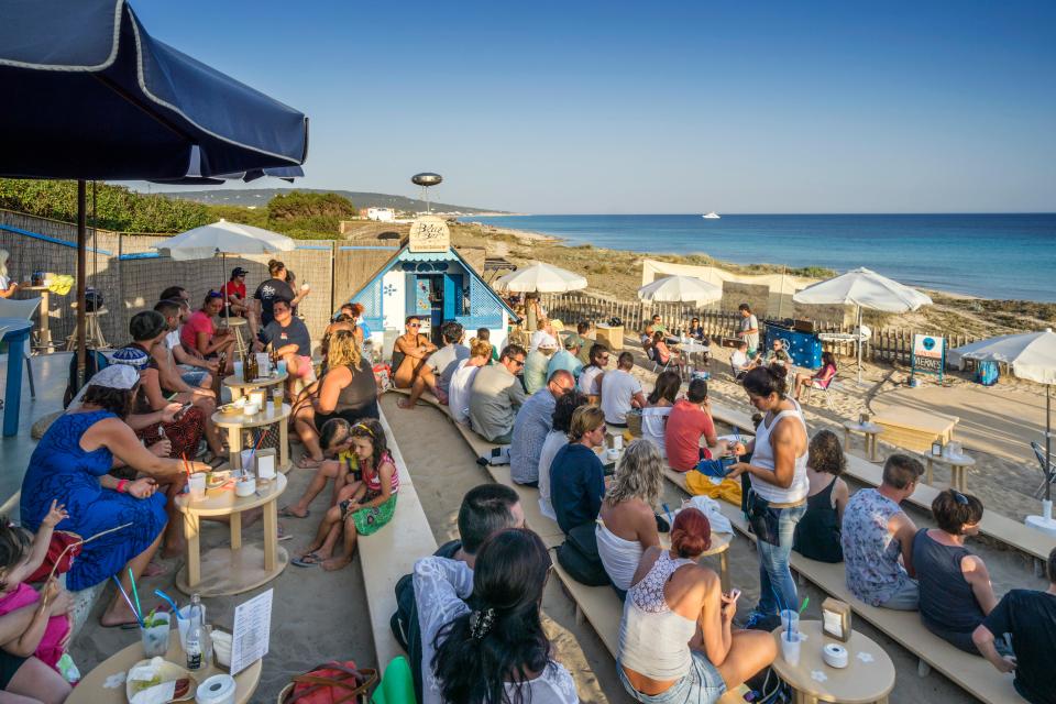Beach bar patrons seated at tables on a sandy beach, overlooking the ocean.