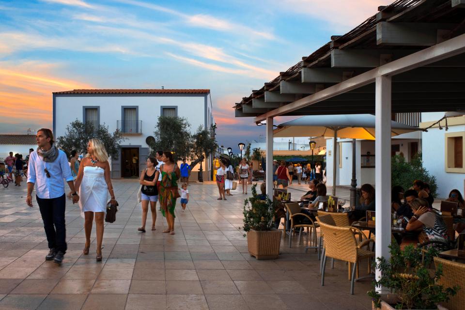 Tourists and locals at an outdoor cafe in a town square at sunset.