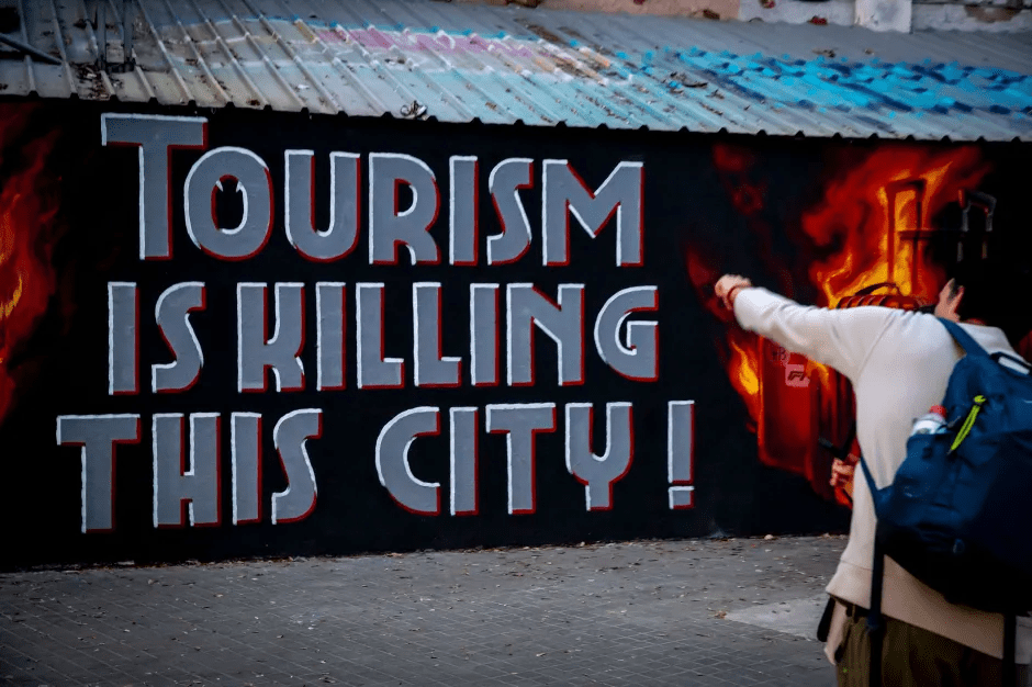 Graffiti depicting "Tourism is killing this city!" with a person pointing at a burning suitcase.