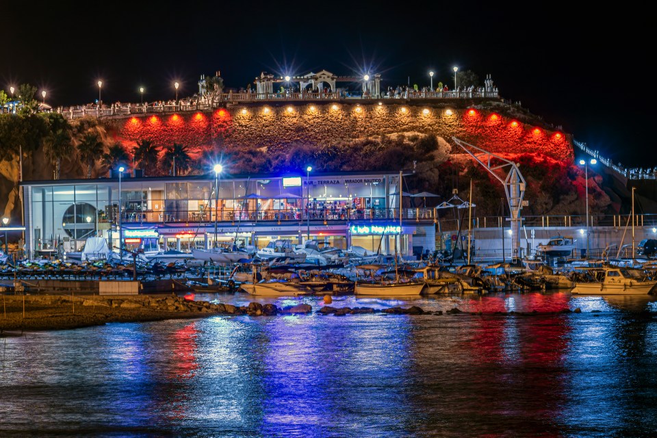 Night view of a harbor with boats and a cliffside restaurant illuminated in red and blue.