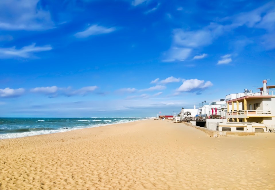 Sandy beach in Faro, Portugal, with ocean waves and buildings in the background.
