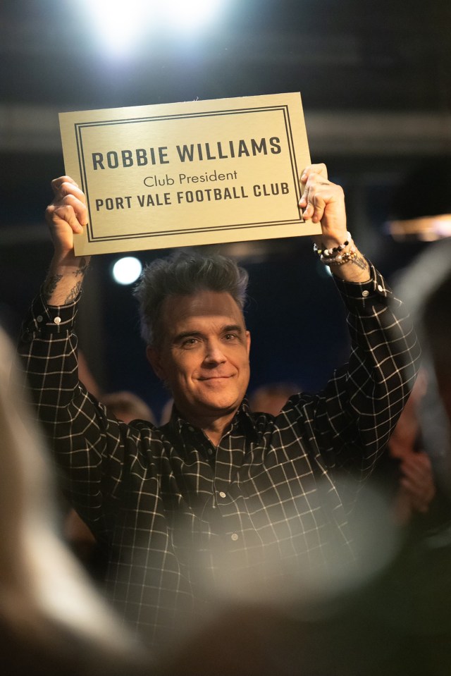 Robbie Williams holding a sign that says "Robbie Williams, Club President, Port Vale Football Club".