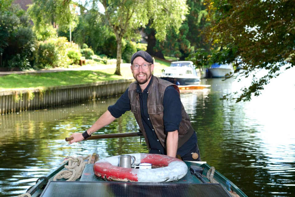 Man in leather vest steering a narrowboat on a canal.