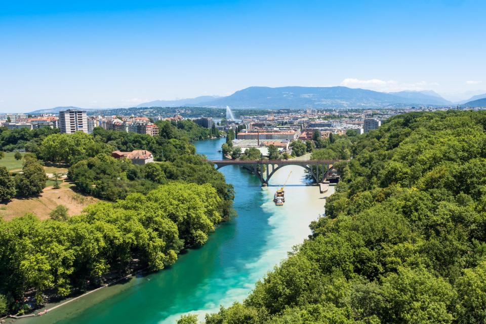 Aerial view of Geneva, Switzerland, showing a river flowing through the city, a bridge, and surrounding greenery.