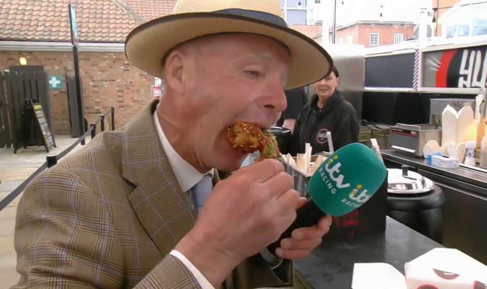 Man in a hat eating a piece of fried chicken while being interviewed.