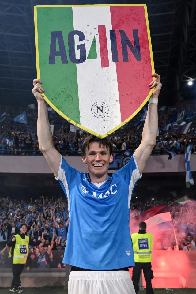 Scott McTominay of Napoli celebrating after a Serie A match, holding a shield that says "AGAIN" in Italian colors.
