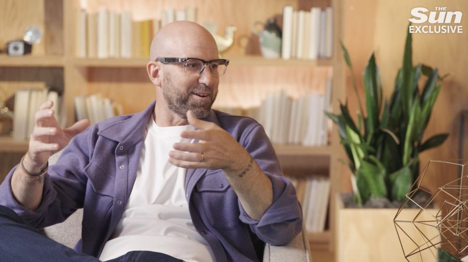 a man wearing glasses and a purple shirt is sitting in front of a bookshelf with the sun exclusive written on it