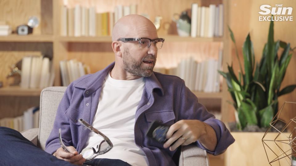a man wearing glasses sits in a chair with the sun exclusive written above him