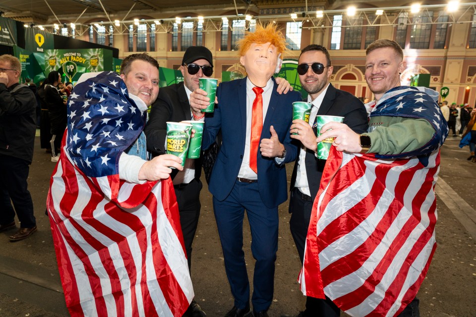 A man dressed as Donald Trump with three other men draped in American flags at a darts event.