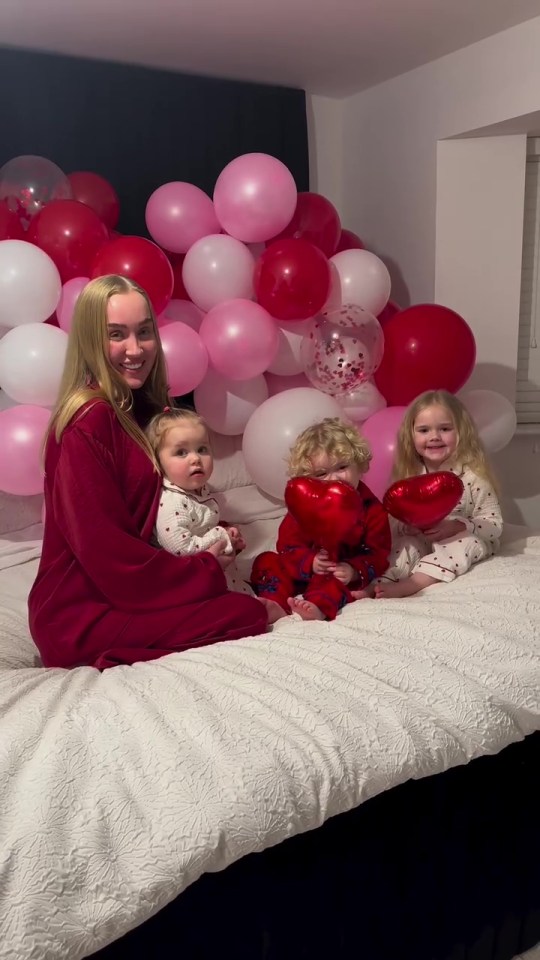 Woman with three young children on a bed with pink and red balloons.