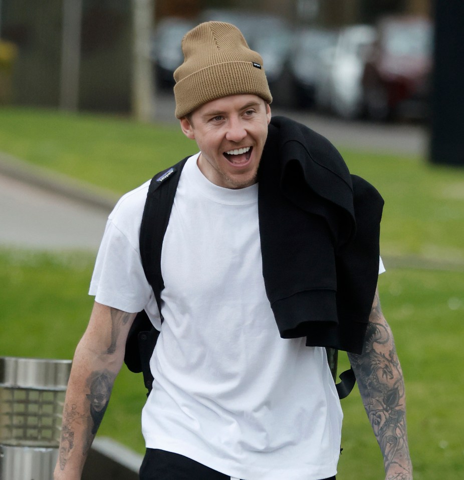 Danny Jones smiling while wearing a tan beanie, white t-shirt, and black jacket.