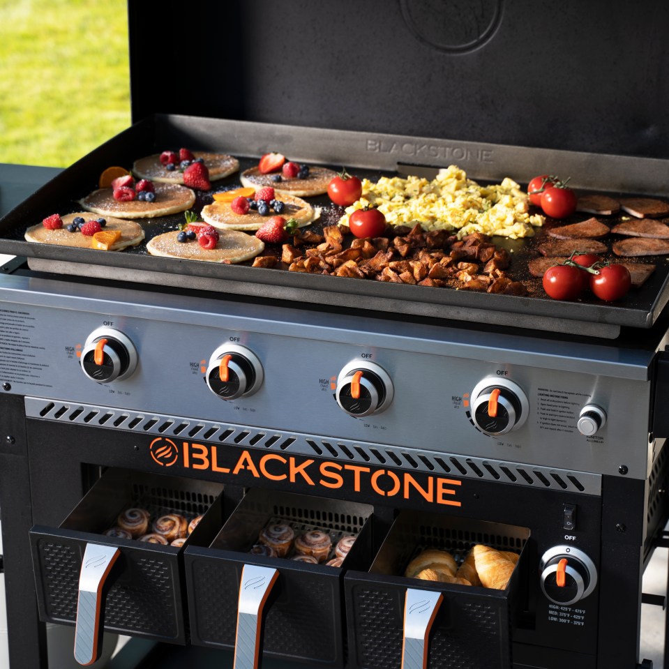 Blackstone griddle cooking breakfast: pancakes with berries, scrambled eggs, potatoes, and bacon.  The griddle has warming drawers with cinnamon rolls and croissants.