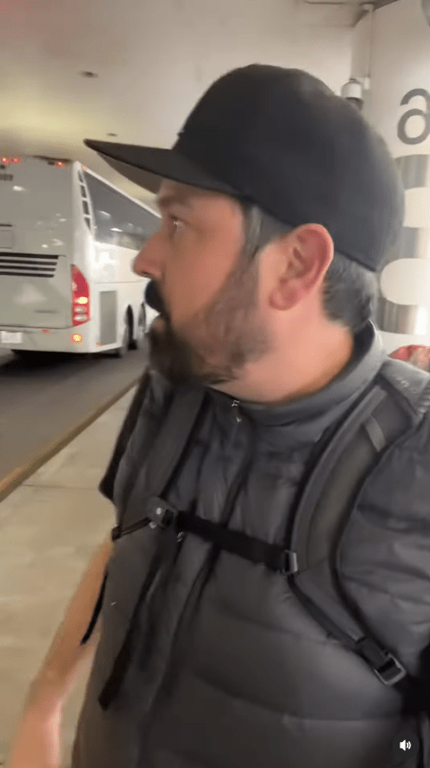 Man in a baseball cap and vest walking past a bus.