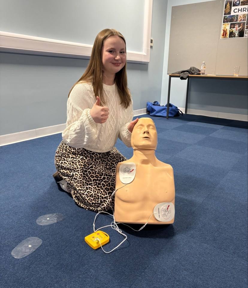 Teenage girl giving a thumbs-up next to a CPR training dummy.